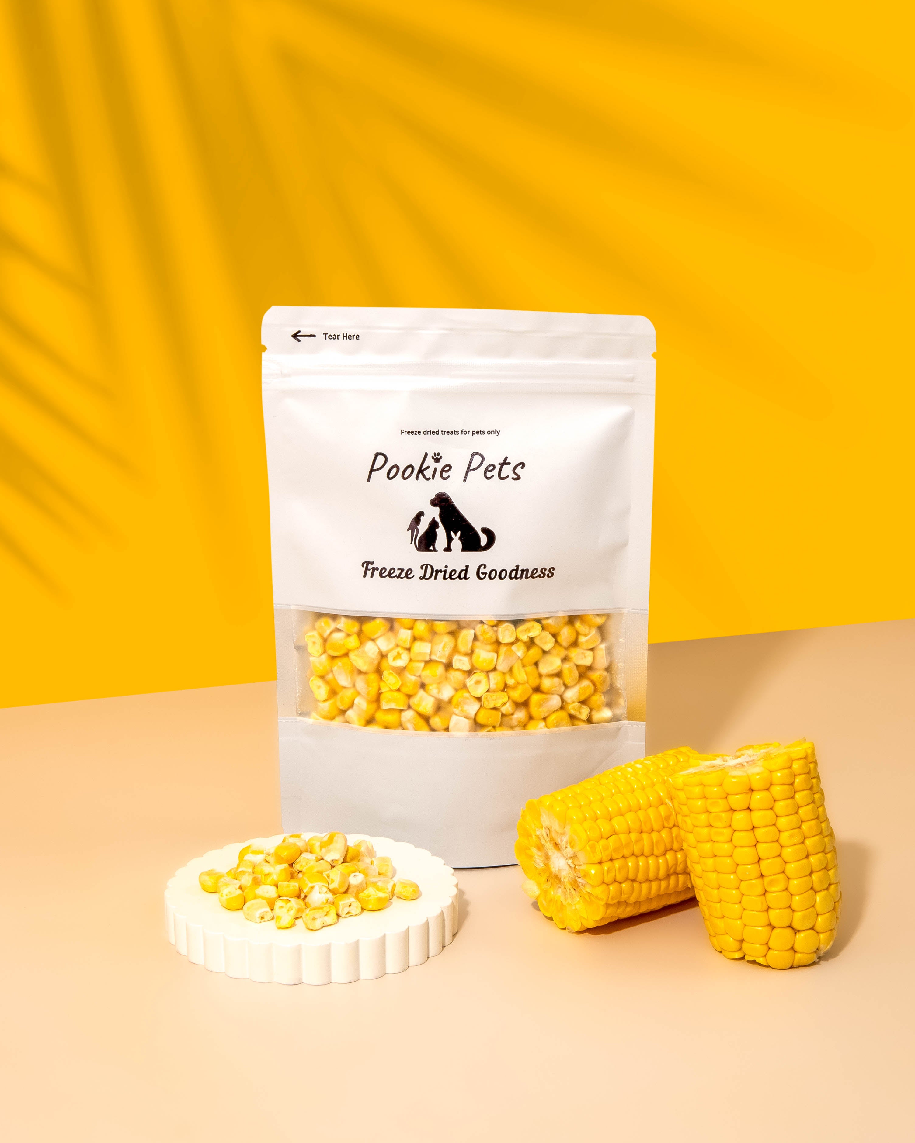 A package of Pookie Pets Freeze-Dried Corn Kernels with a transparent window showing the dried corn inside. The package is displayed alongside fresh corn cobs and a dish with scattered corn kernels.