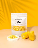 A package of Pookie Pets Freeze-Dried Corn Kernels with a transparent window showing the dried corn inside. The package is displayed alongside fresh corn cobs and a dish with scattered corn kernels.